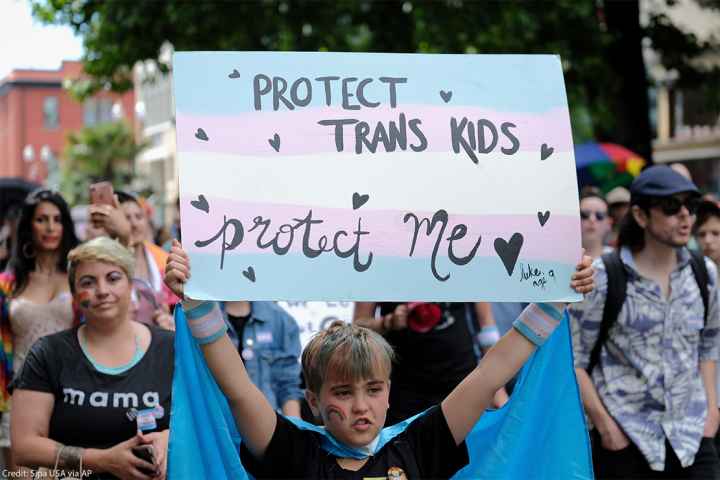 A young child holds up a sign reading "PROTECT TRANS KIDS, PROTECT ME - luke, age 9" at a Trans Pride March.