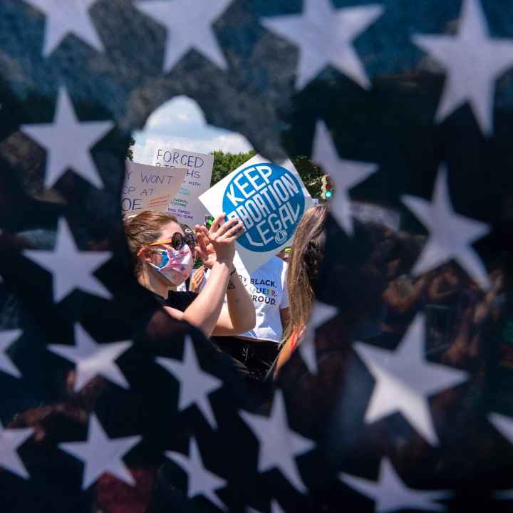 Abortion rights activists are seen through a hole in an American flag as they protest outside the Supreme Court in Washington, Saturday, June 25, 2022.