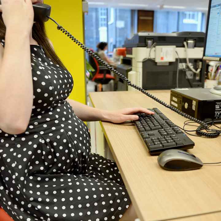 A pregnant worker using an office phone while typing on a keyboard.